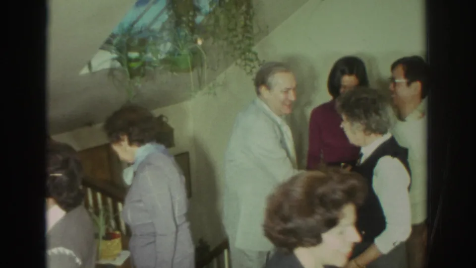 1972 Toronto home-movie frame showing adults in a private interior near a staircase, talking and greeting one another.