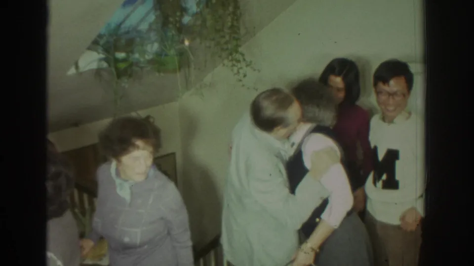 1972 Toronto frame showing adults greeting with a hug as other guests stand nearby in a private home setting.