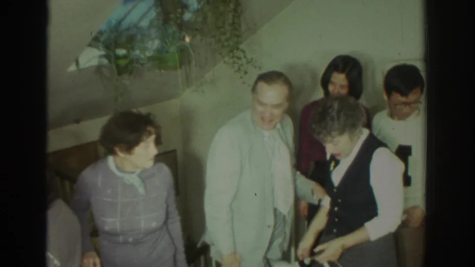 1972 Toronto home-movie frame of adults in sweaters and vests conversing near a stair landing inside a private residence.