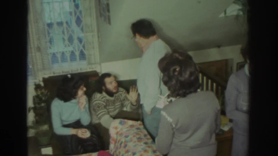 1972 Toronto home-movie frame showing adults talking around a small table, with a curtained window and security grille behind them.