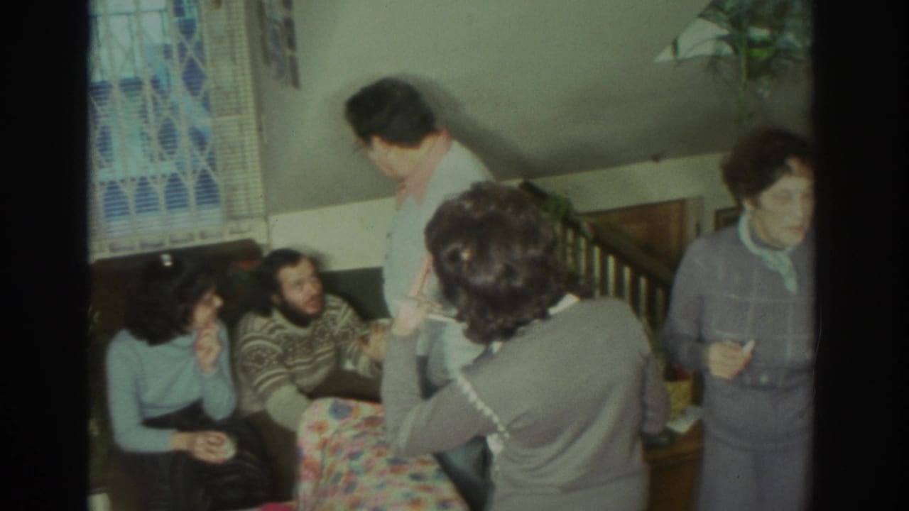 1972 Toronto home-movie frame showing adults in sweaters and cardigans gathered in a private living room around a floral couch.