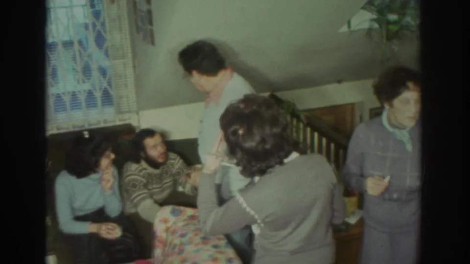 1972 Toronto home-movie frame showing adults in sweaters and cardigans gathered in a private living room around a floral couch.