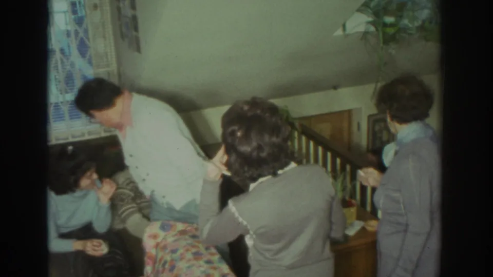 1972 Toronto home-movie frame with a man in a light cardigan leaning toward relatives seated beside a floral couch.