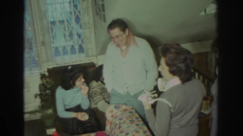 1972 Toronto home-movie frame showing a man in glasses standing near seated relatives, with lace curtains and houseplants behind them.