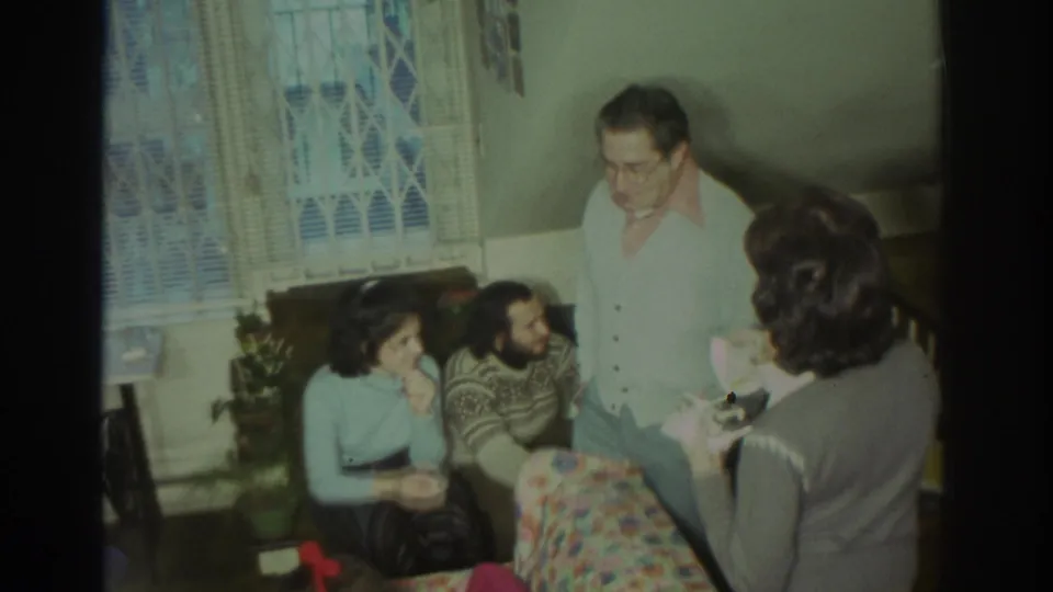 1972 Toronto home-movie frame of adults talking in a private living room with a floral couch, lace-curtained window, plants, and stair railing.