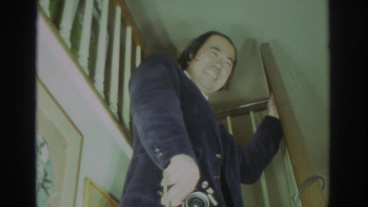 In a 1972 Toronto home-movie frame, a smiling man stands on a wood staircase holding a still camera and looking downward.