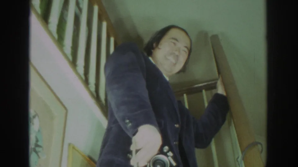 In a 1972 Toronto home-movie frame, a smiling man stands on a wood staircase holding a still camera and looking downward.