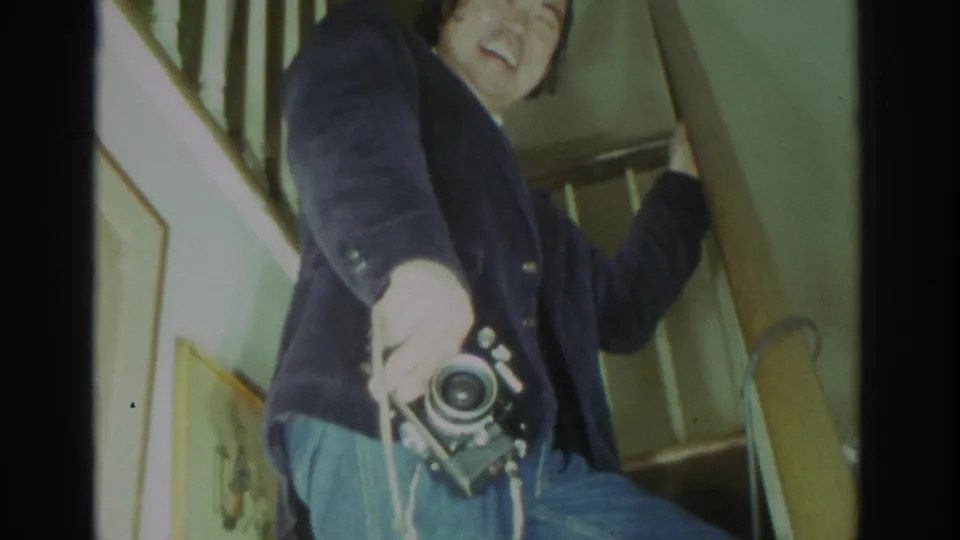 A low-angle 8mm frame shows the man on the staircase, partly cropped, holding a camera in a private Toronto interior.