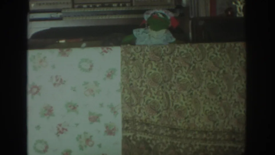 1972 Toronto home interior frame with the green puppet near a white bonnet-like cloth, stereo equipment, and patterned upholstery.