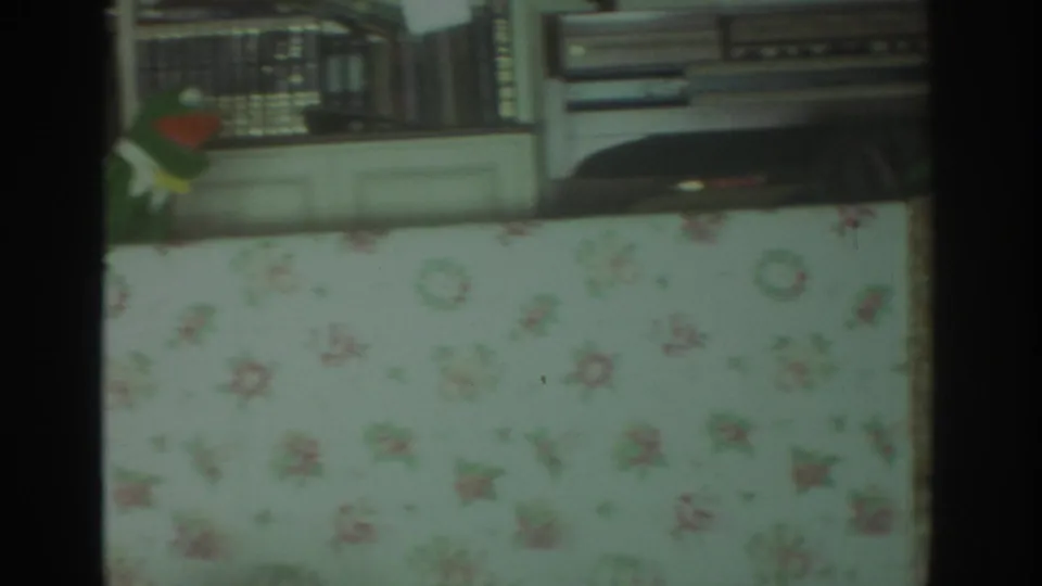 1972 Toronto home-movie frame with a floral curtain foregrounding a domestic shelf area with books and stereo equipment.