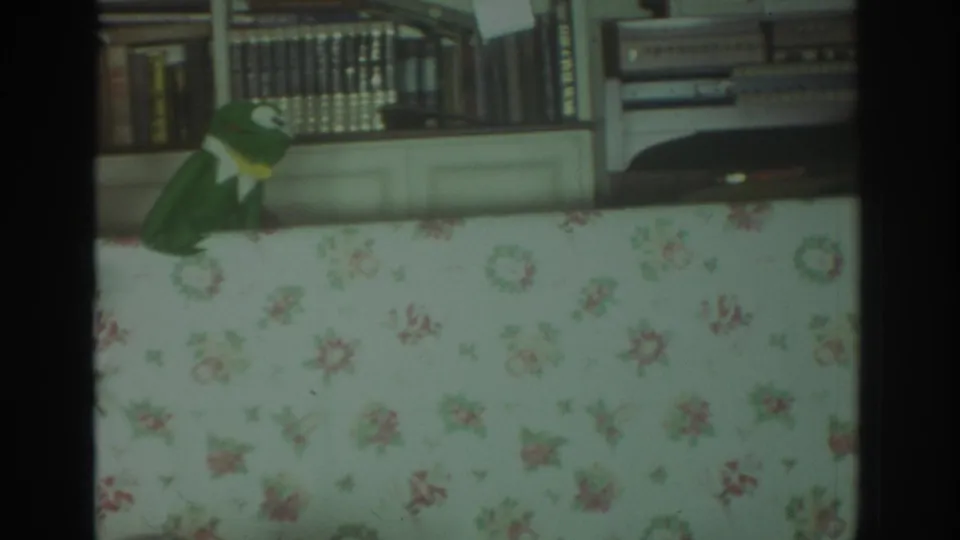 1972 Toronto home-movie frame showing a green frog puppet leaning over a floral curtain in a private living room.