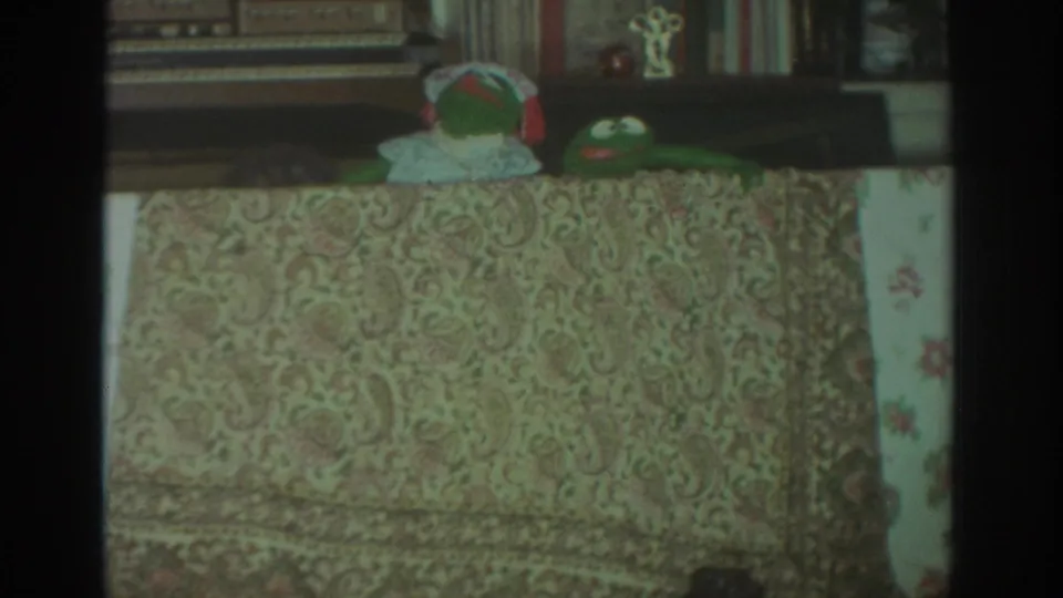 1972 Toronto home-movie frame with a bonneted handmade puppet and green frog puppet visible behind a paisley table covering in a private living room.