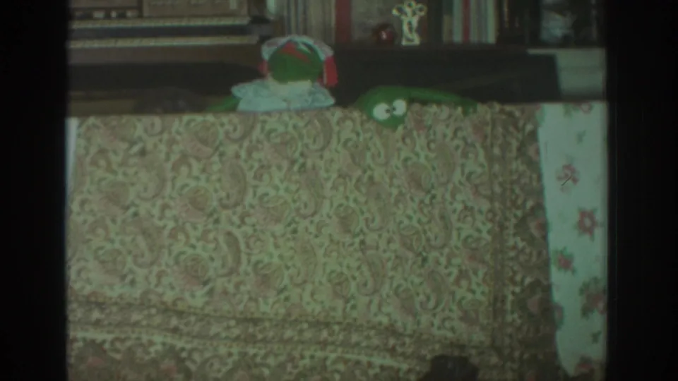 1972 Toronto 8mm frame with a green frog puppet peeking over a paisley tablecloth in a private living-room interior.