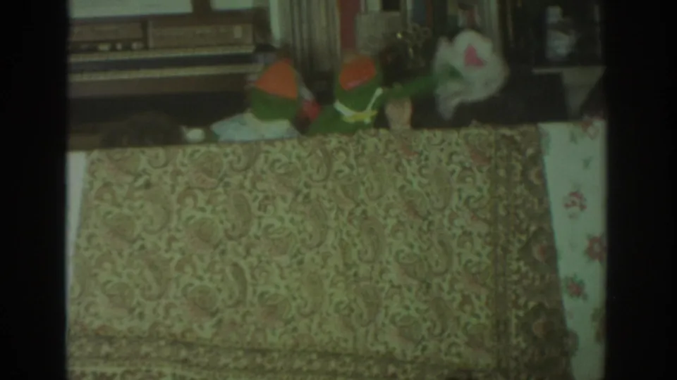 1972 Toronto 8mm frame showing colorful hand puppets performing behind a patterned tablecloth in a domestic interior.