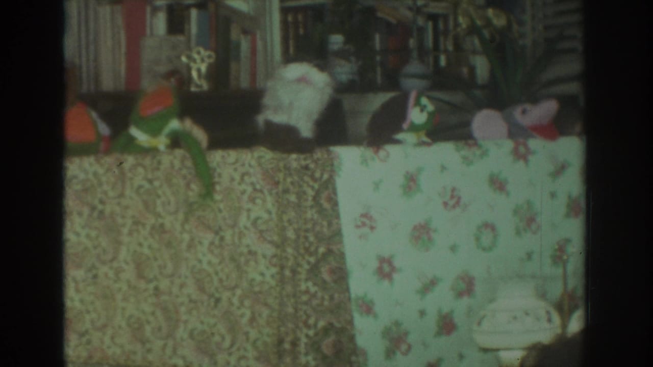 Several puppets are arranged behind a floral table covering with shelves and a small trophy-like object behind them.