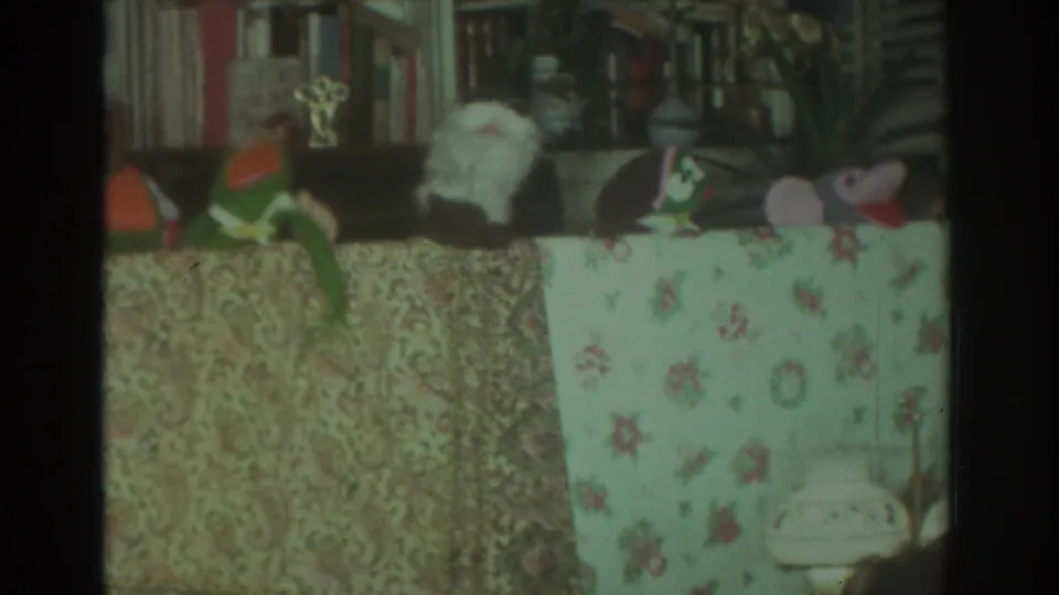 1972 Toronto home-movie frame with multiple hand puppets behind a floral tablecloth and shelves in the background.
