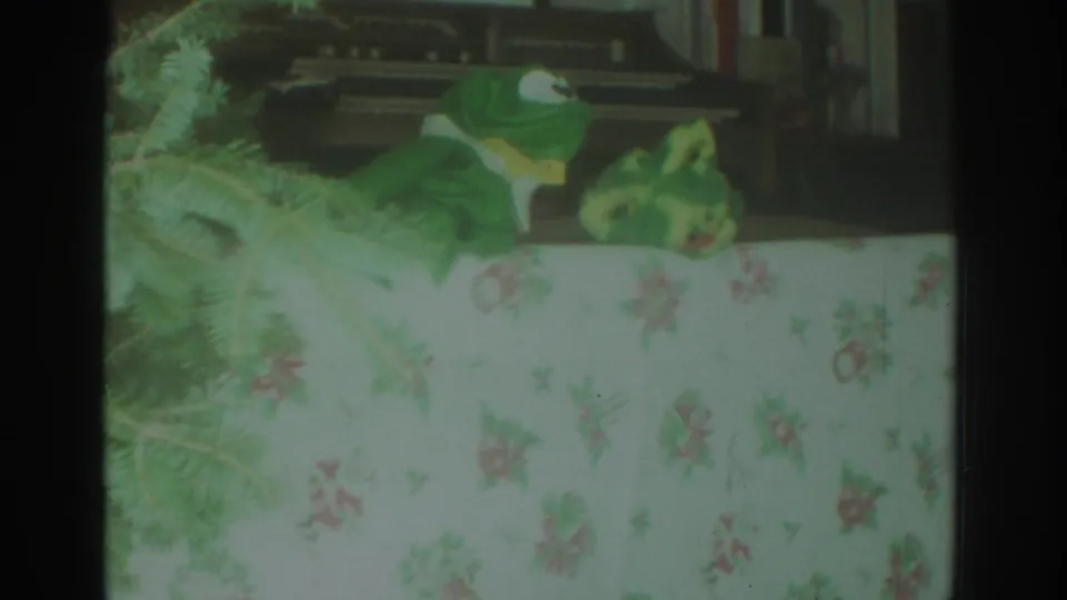 1972 Toronto 8mm frame showing a green frog puppet with a yellow bowtie beside another puppet over a floral tablecloth.