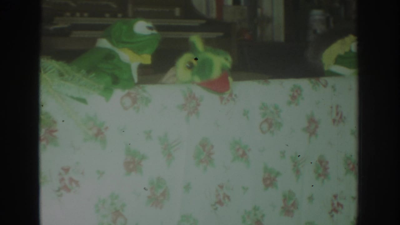 1972 Toronto 8mm frame showing two green puppets behind a pale floral tablecloth, with dark film borders and a soft indoor background.