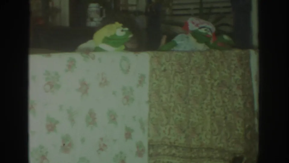 1972 Toronto 8mm frame showing a green puppet and a patterned puppet figure behind floral and paisley table coverings.