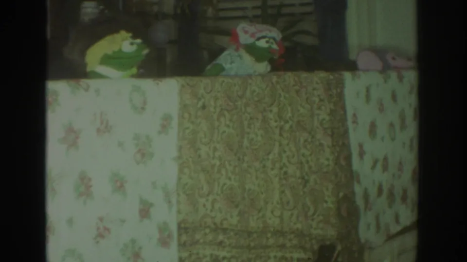 1972 Toronto 8mm frame with a green puppet, patterned puppet figure, pale floral cloth, paisley fabric, and small pink object at right.