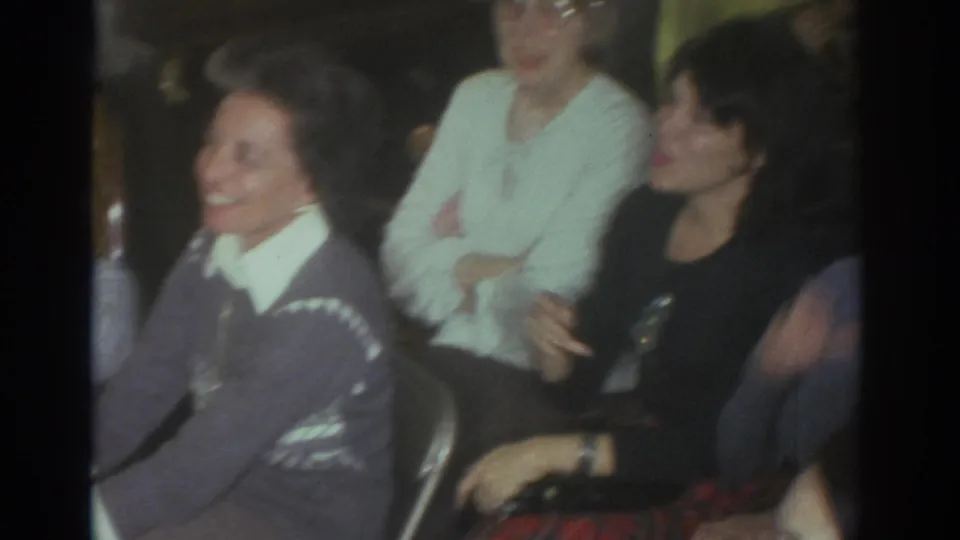 Several adults sit close together in the 1972 Toronto living room, including women smiling and laughing during the puppet-show moment.