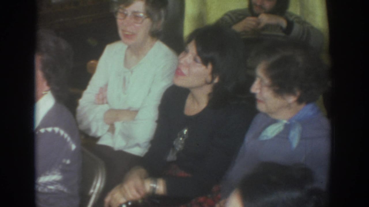 Adults seated indoors, including women in sweaters and a person turned away in the foreground.