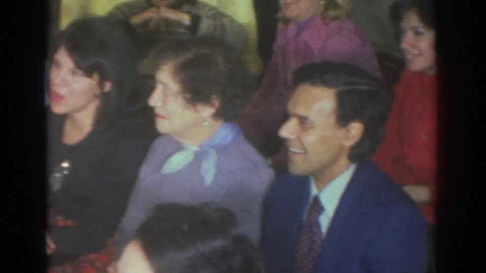 Toronto 1972 frame with adults smiling at an indoor gathering, including a man in a blue suit and women in bright tops.