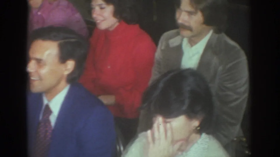 Toronto 1972 home-movie frame showing a smiling man in a blue suit beside adults seated in a crowded private interior.