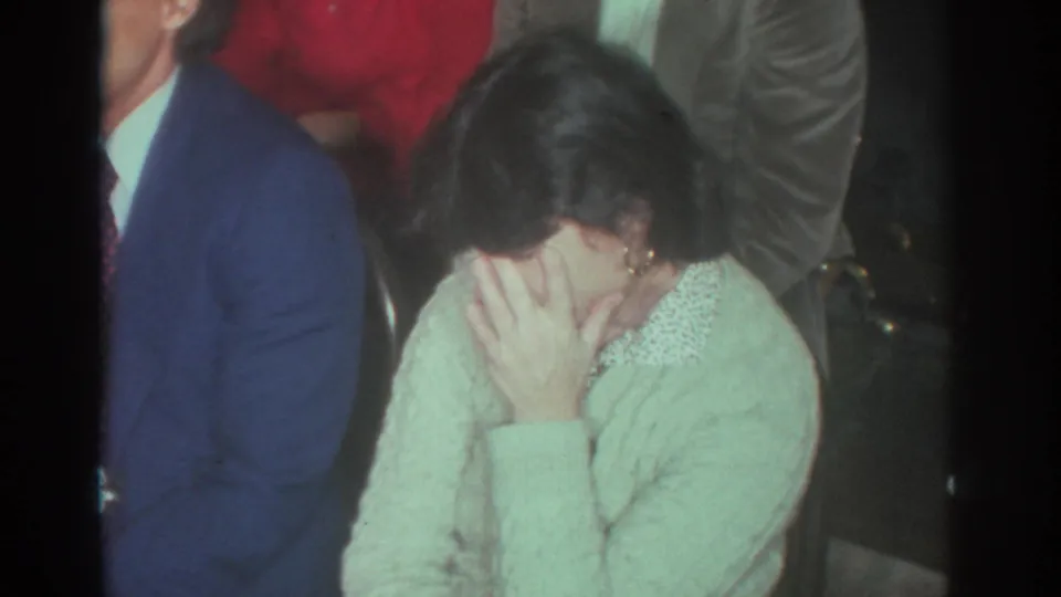 Toronto 1972 frame of a woman covering her face in amusement or shyness during a crowded indoor home-movie gathering.