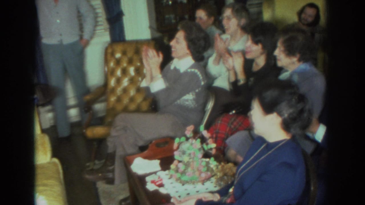 Adults seated in a living room clap near a decorated cake.