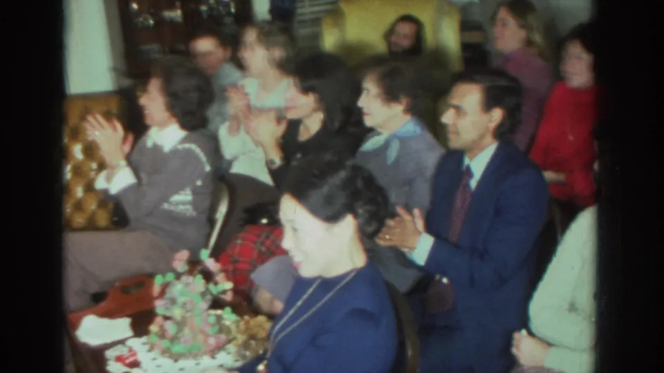 A wider 1972 Toronto home-movie frame shows adults applauding in a crowded living room with a cake on the table.