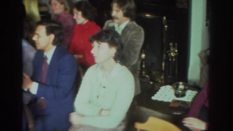 At the Toronto gathering, the woman in a pale sweater sits with arms crossed while nearby adults continue clapping in the living room.