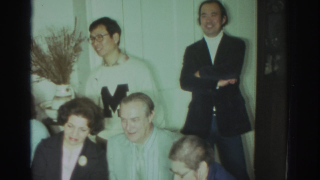 Adults seated and standing indoors, including a man in a white lettered sweater and a woman in a dark blazer.