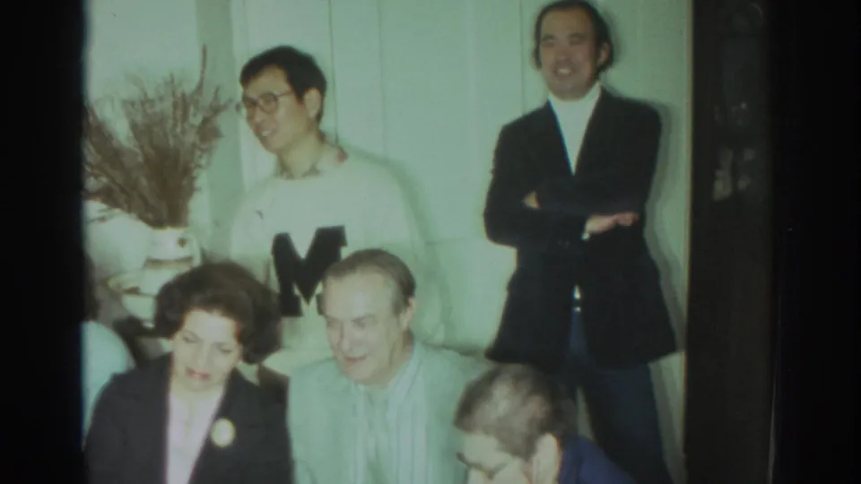 1972 Toronto home-movie frame of adults gathered indoors, with a man in a white sweater, a seated woman in a dark blazer, and pale wall paneling.