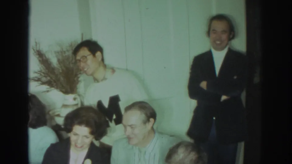 1972 Toronto indoor gathering with adults seated and standing, including a white sweater with a large letter and a dark jacket over a turtleneck.