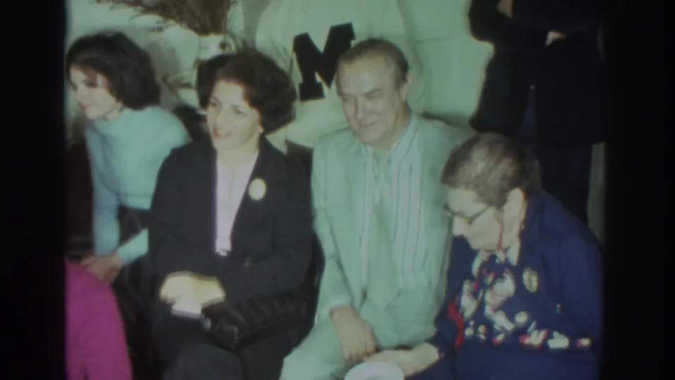 1972 Toronto social-gathering frame with seated adults, a woman in a dark blazer, an older woman in glasses, and men standing behind.