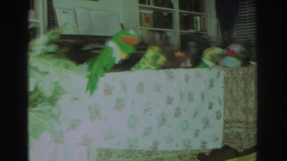 1972 Toronto home-movie frame of a green frog puppet and small toy figures arranged on a floral-covered table in a living room.