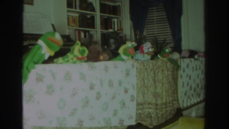1972 Toronto home-movie frame with a frog puppet at left and smaller figures lined across a cloth-covered table in a living room.