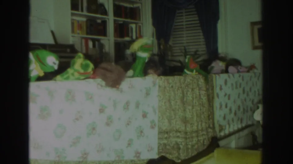 1972 Toronto home-movie frame showing multiple puppets on a floral-covered table, with bookshelves, blinds, and dark curtains in the background.