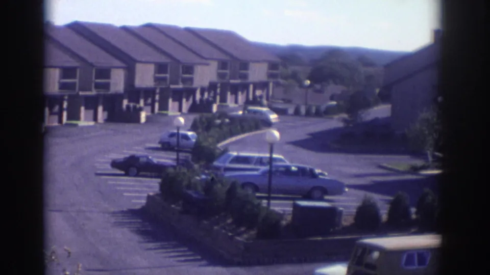 1972 Toronto residential complex with matching townhouses, a paved lot, parked cars, and distant wooded hills.