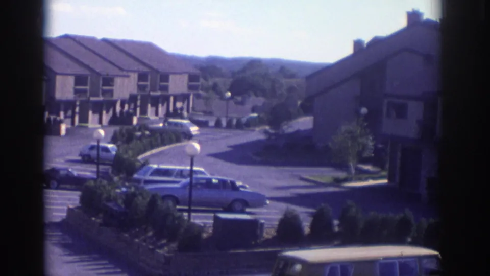 Home-movie frame from 1972 Toronto showing the residential complex as the camera pans across cars, shrubs, and townhouse facades.