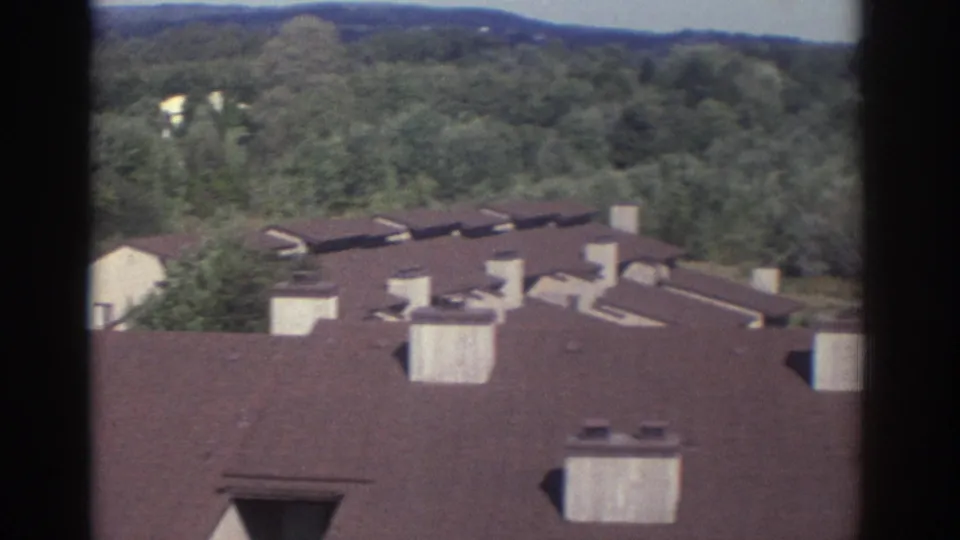 1972 Toronto home-movie view over apartment or townhouse rooftops, chimneys, roof vents, and a green hillside beyond.