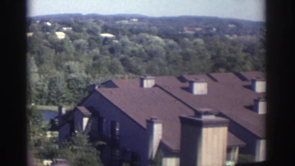 Soft 8mm view of 1972 Toronto residential roofs, chimneys, dense trees, distant houses, and low wooded hills.
