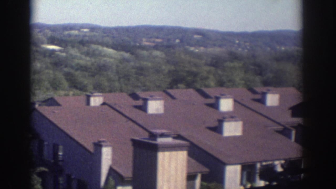 1972 Toronto home-movie frame showing brown-roofed suburban apartment or townhouse buildings below a tree-covered hillside, partially framed by dark window edges.