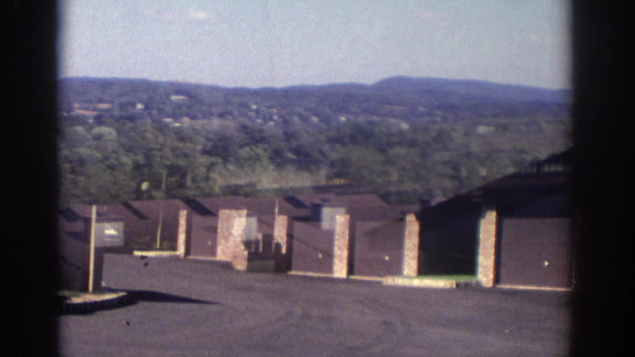 1972 Toronto residential complex seen from above, with brown roofs, garage bays, brick sections, paved driveways, and wooded hills in the background.