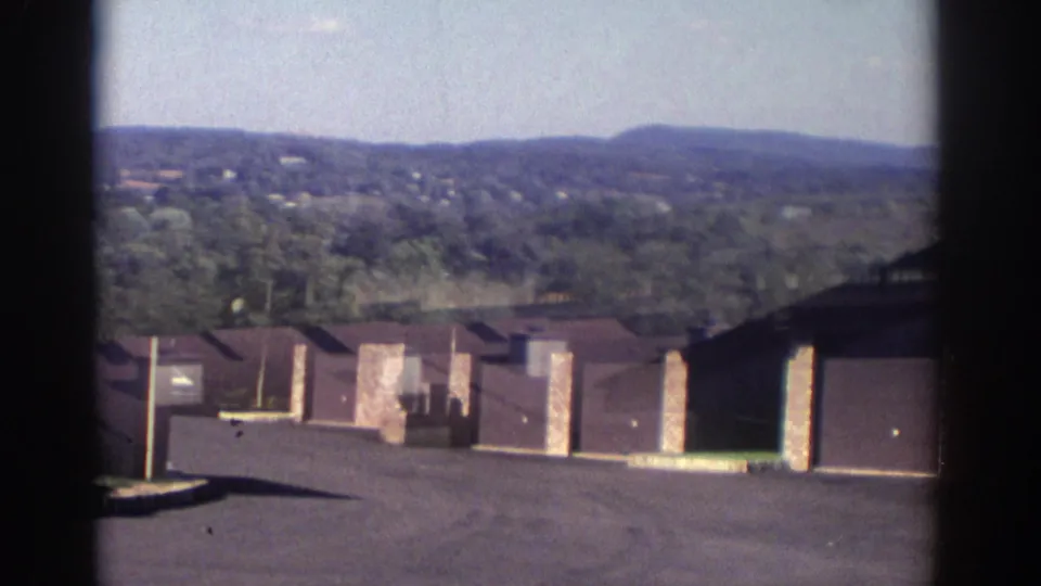 1972 Toronto residential complex seen from above, with brown roofs, garage bays, brick sections, paved driveways, and wooded hills in the background.