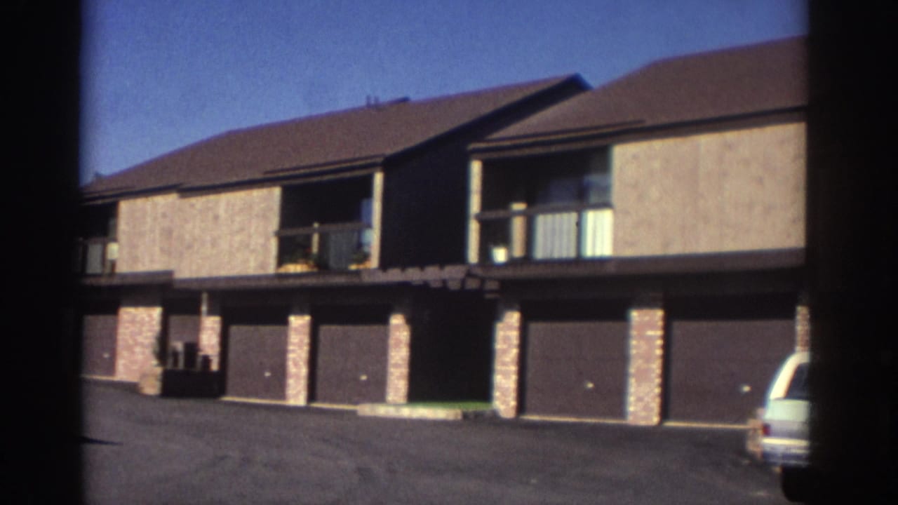 1972 Toronto home-movie view of a townhouse-style apartment facade with stucco walls, brick supports, balconies, attached garages, and a light-colored station wagon partly visible.