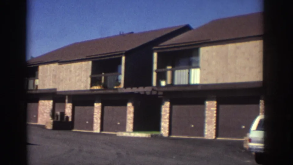 1972 Toronto home-movie view of a townhouse-style apartment facade with stucco walls, brick supports, balconies, attached garages, and a light-colored station wagon partly visible.