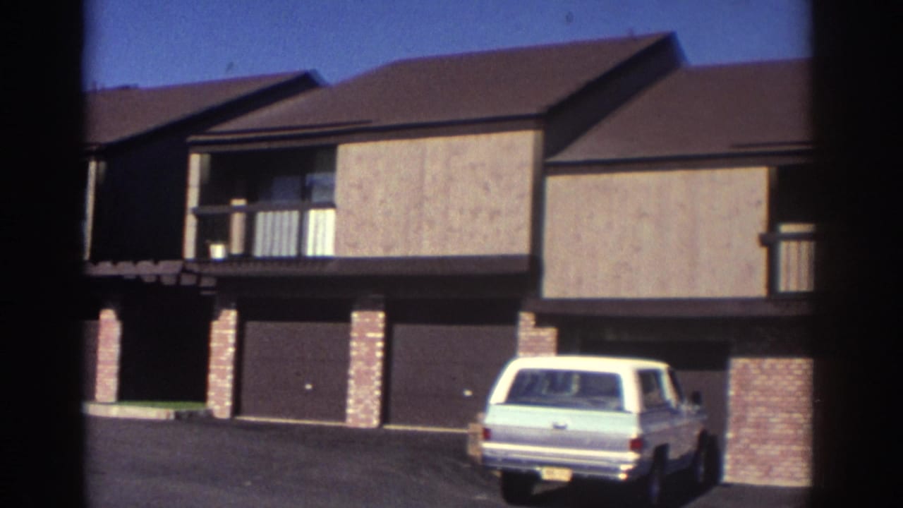 1972 Toronto residential exterior showing attached garage doors, brick columns, upper balconies, a paved driveway, and a light-colored vintage station wagon parked near the building.