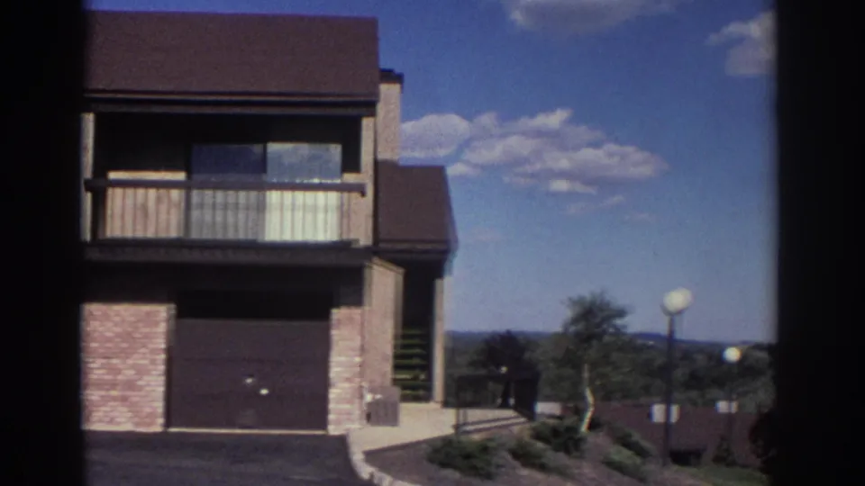 1972 Toronto 8mm frame as the camera pans past the housing estate toward shrubs, walkway, lampposts, and distant low hills or treeline.